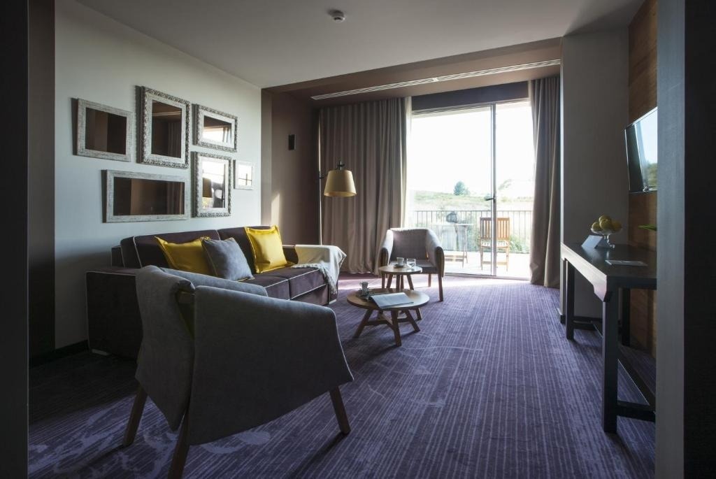 Un salón moderno y acogedor con alfombra morada, un sofá oscuro con cojines amarillos y grises, y una gran puerta corrediza de cristal que da a un balcón.