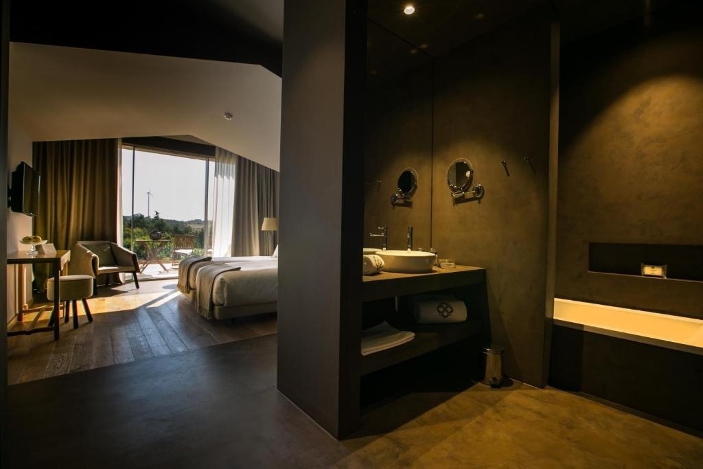 Una habitación de hotel moderna y elegante de planta abierta, que conecta un dormitorio luminoso con vistas panorámicas y un baño adyacente de tonos oscuros.