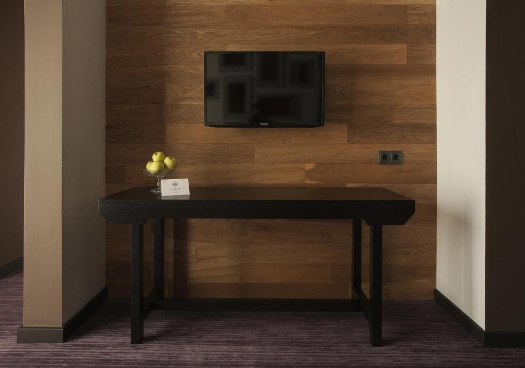 Una pared de madera con un televisor montado y un escritorio negro debajo con un tazón de manzanas y una tarjeta, sobre una alfombra morada.