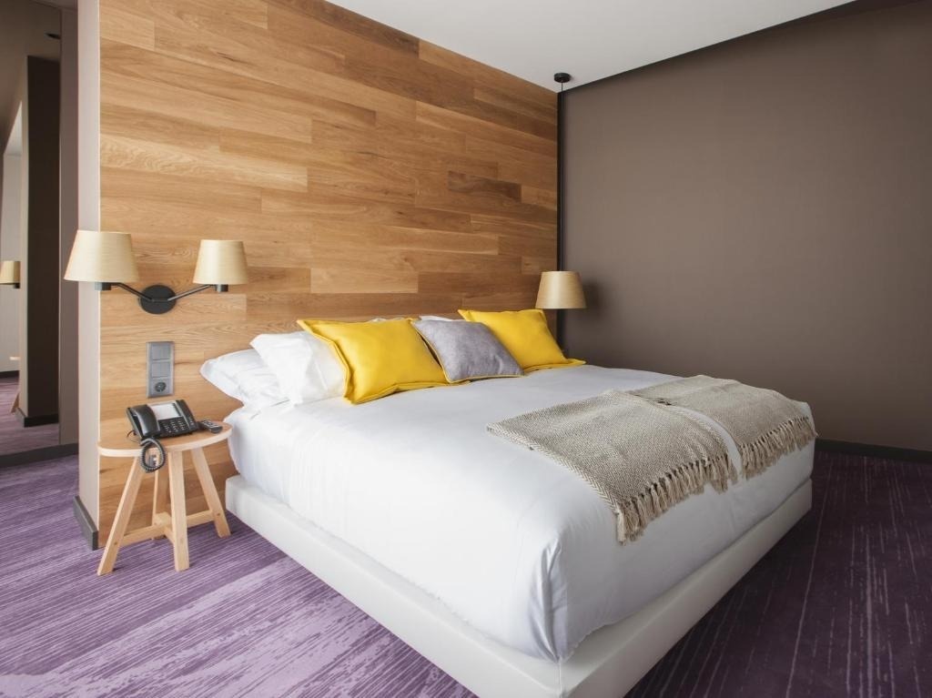 Un moderno dormitorio presenta una cama con cabecero de madera, almohadas amarillas y grises, y una alfombra morada con diseño.