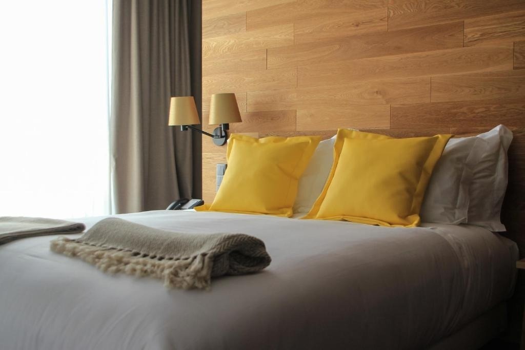 Una cama de hotel está hecha con sábanas blancas, almohadas amarillas y un cabecero de madera.