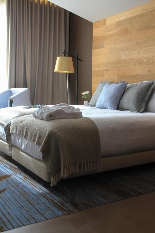 Una habitación moderna presenta una cama con ropa blanca, almohadas azules y grises, y una manta, junto a una lámpara de pie, una pared de madera y una alfombra de patrones azules.