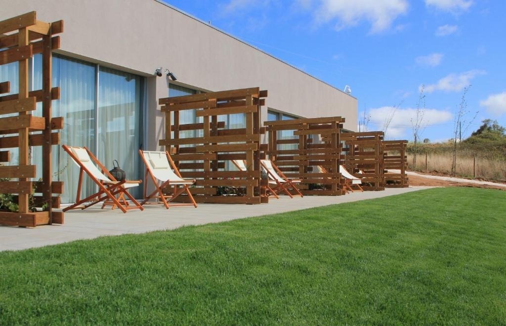 Una fila de patios individuales, cada uno con divisiones de madera y tumbonas blancas, se extiende a lo largo de un edificio moderno, con un césped verde en primer plano y un cielo azul con nubes.