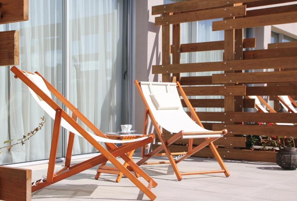 En una terraza luminosa, dos sillas plegables de madera y lona blanca con una pequeña mesa con una taza se ubican junto a una celosía de madera, con puertas de cristal y cortinas claras al fondo.