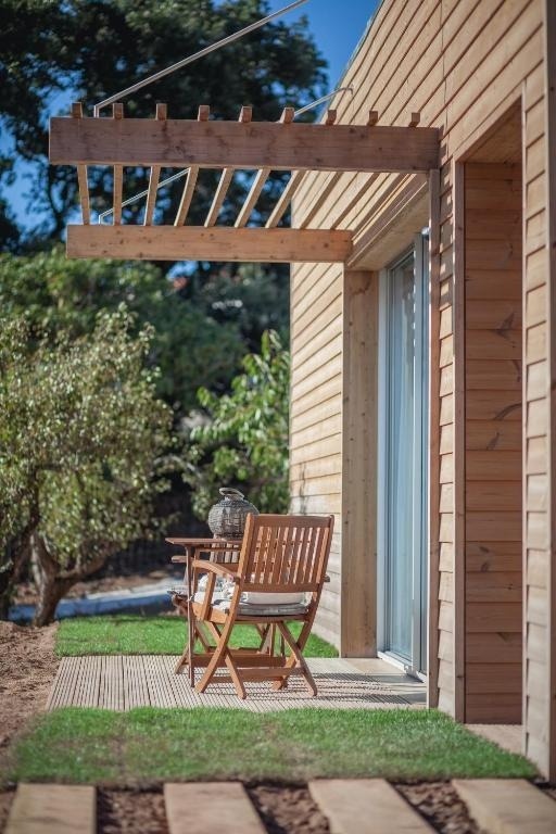 Vista exterior de una casa moderna de madera con una pérgola, un patio con mobiliario de jardín y un sendero de césped con listones de madera.
