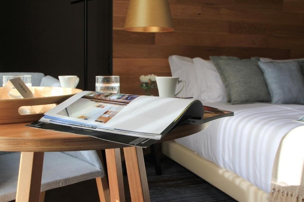 Una acogedora habitación de hotel muestra una cama hecha, una mesita con una revista abierta, un vaso de agua y una taza de café, con una pared de madera de fondo.