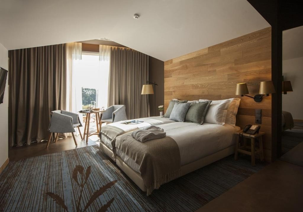 Una habitación de hotel luminosa presenta una cama grande con cabecero de madera, una zona de estar junto a una ventana con cortinas y una alfombra estampada.