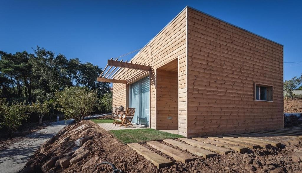 Una casa moderna de revestimiento de madera, con un pequeño patio y pérgola, se asienta en un entorno rural con vegetación y un sendero de tablones de madera bajo un cielo azul claro.