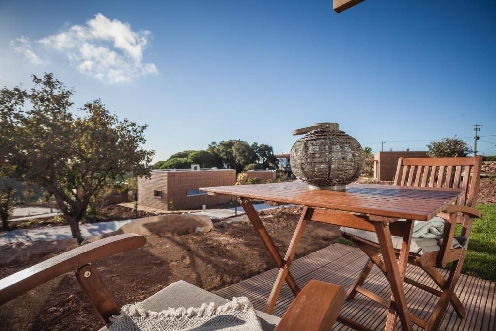 Una terraza de madera con una mesa y sillas, decorada con una linterna, ofrece vistas a edificios modernos y árboles bajo un cielo azul despejado.