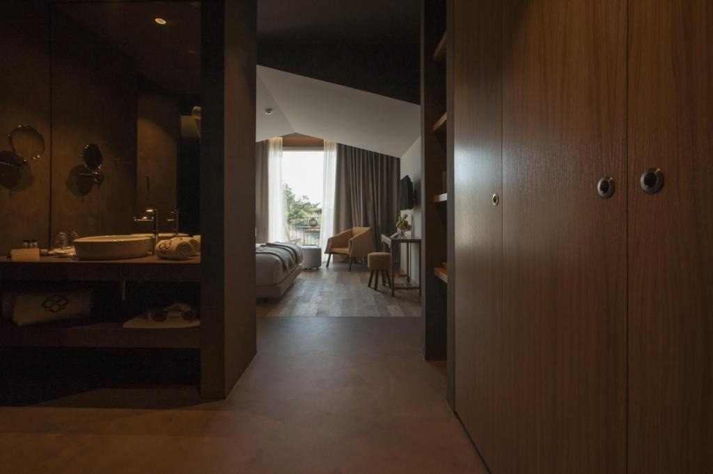 La imagen muestra el interior de una moderna suite de hotel, con un baño a la izquierda, un dormitorio con vista exterior al fondo y armarios de madera a la derecha.