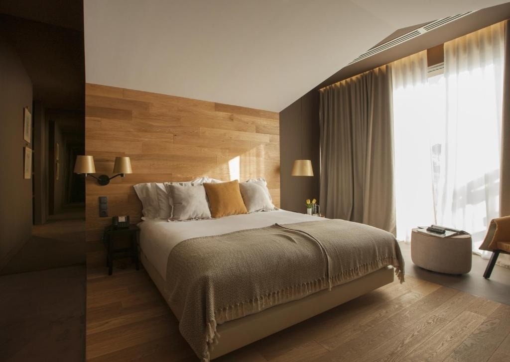 Un moderno dormitorio luminoso presenta una cama con cabecero de madera, almohadas en tonos tierra y grandes ventanales cubiertos por cortinas claras y oscuras.