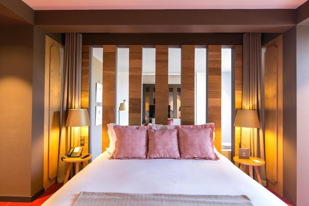 Una cama grande con almohadas rosas y un cabecero de listones de madera que actúa como divisor parcial se presenta en un dormitorio moderno con iluminación cálida.