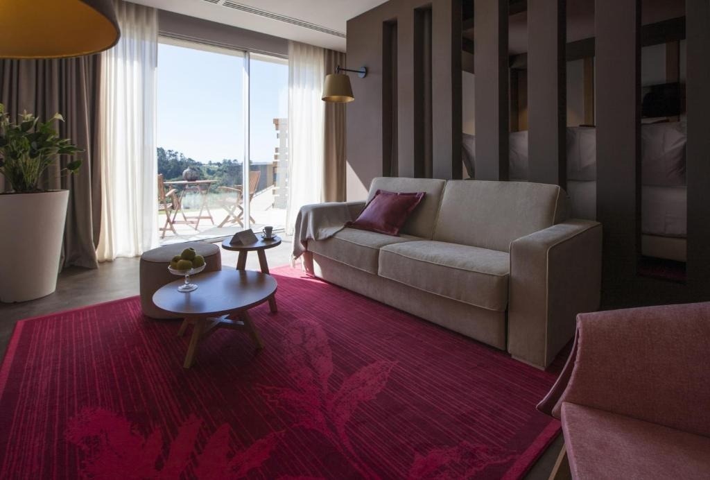 Un luminoso salón moderno con un sofá beige, una alfombra roja con diseño floral, mesas de centro de madera y amplios ventanales con vista a un balcón exterior y vegetación.