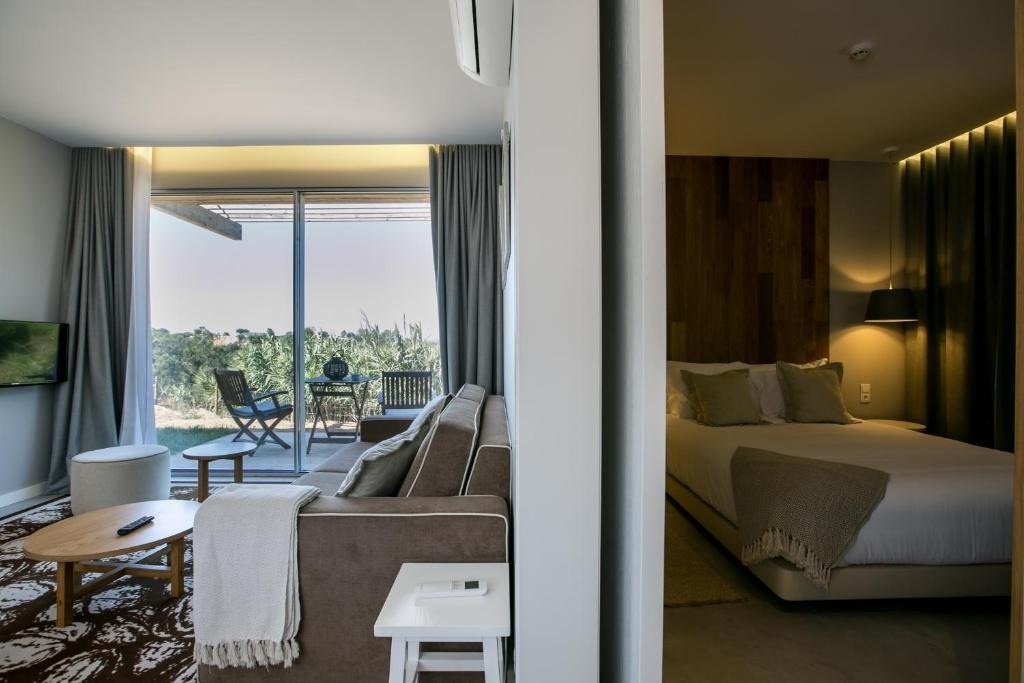 Un interior moderno revela una sala de estar con sofá y un balcón con vistas a la naturaleza, junto a un dormitorio con una cama grande y cabecero de madera.