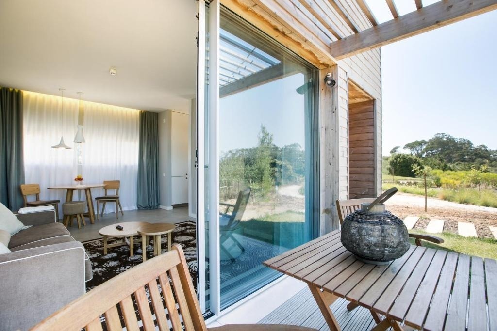 La imagen muestra un espacio interior moderno con sala de estar y comedor, que se abre a un patio exterior de madera con mesa y sillas, con un paisaje natural de árboles y vegetación de fondo.