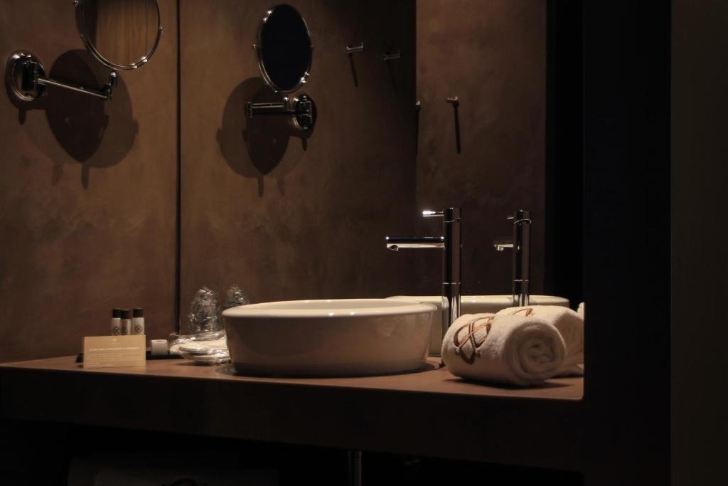 Un baño moderno con iluminación tenue exhibe un lavabo de tazón blanco, grifos elegantes y espejos de aumento sobre una encimera de tonos tierra.