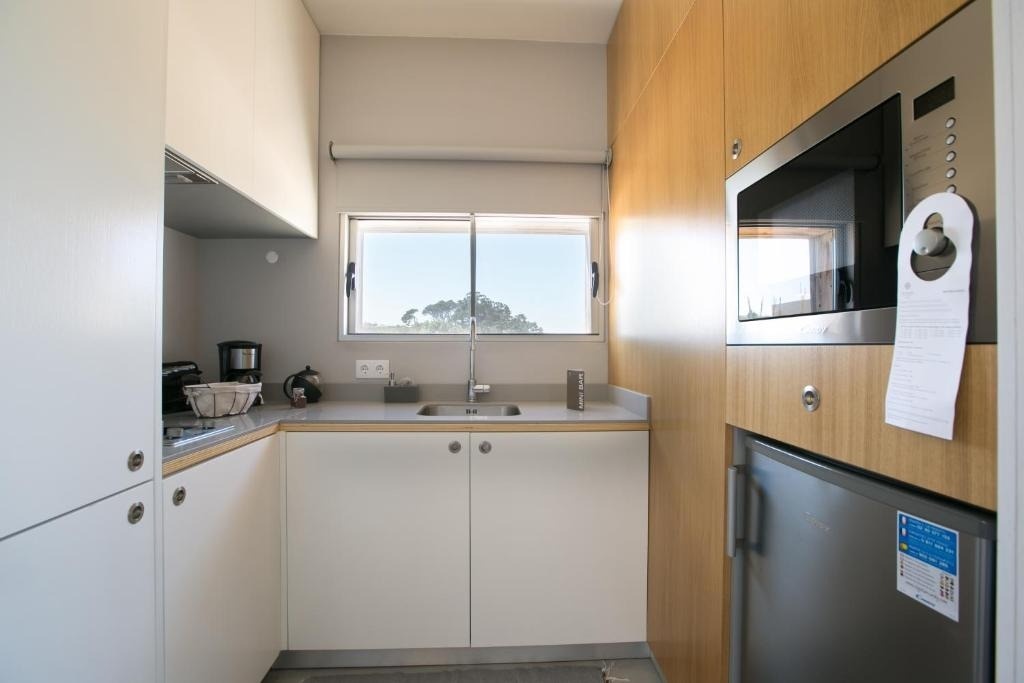 Una moderna y compacta cocineta con gabinetes claros, un fregadero bajo una ventana que ofrece vistas a la naturaleza, y electrodomésticos como un microondas y un mini-refrigerador.