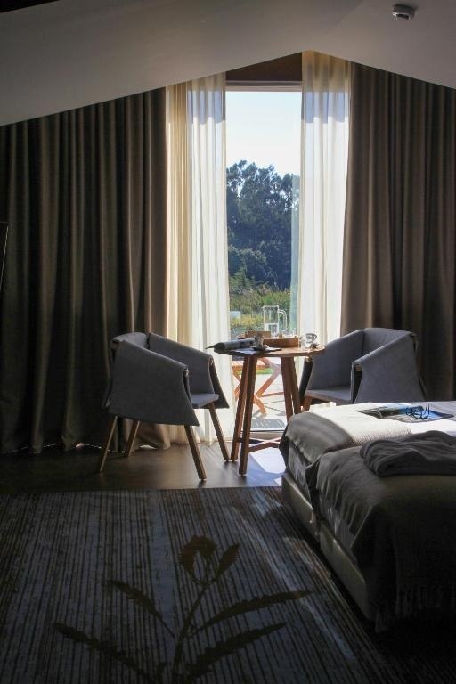 Una habitación de hotel luminosa con una cama, una mesa con dos sillas y una gran ventana que da a un paisaje verde.