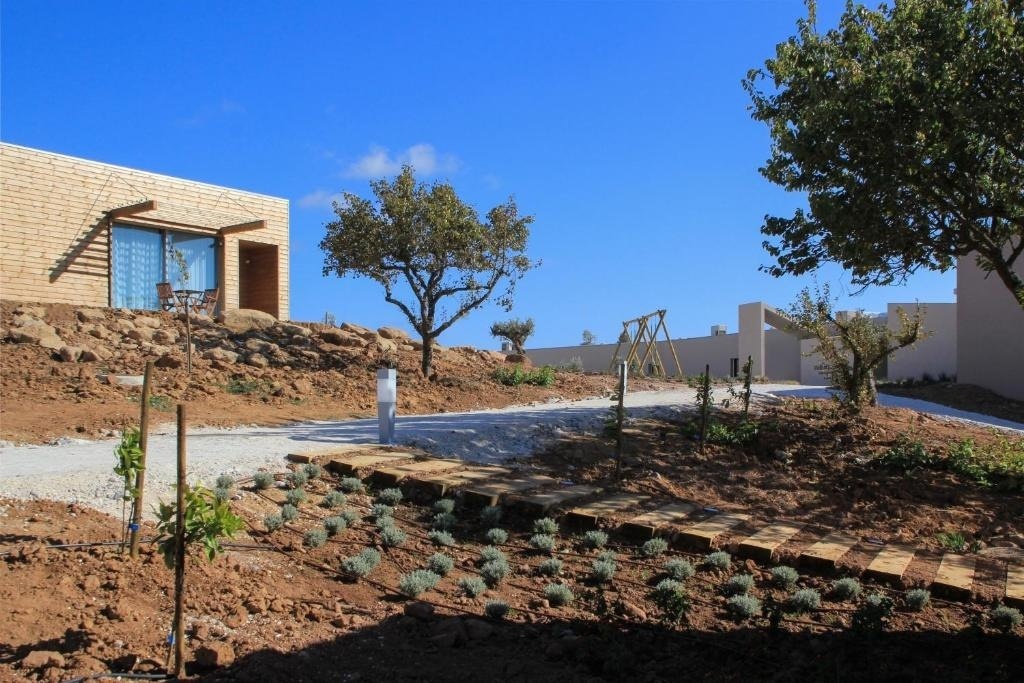 La imagen muestra un complejo de edificios modernos, incluyendo uno con revestimiento de madera, integrado en un paisaje árido y rocoso con vegetación joven, senderos y un columpio de madera, bajo un cielo azul claro.