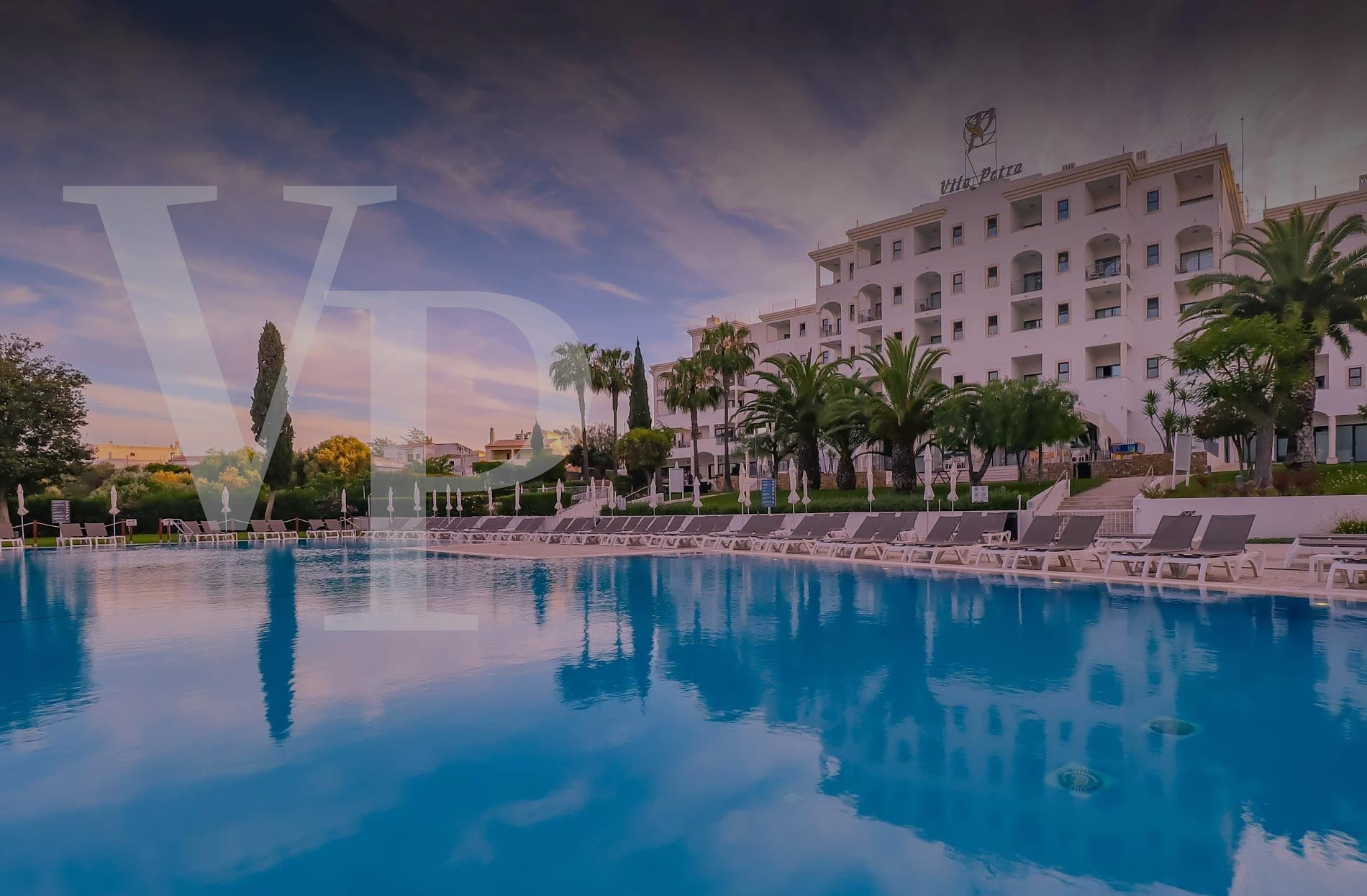 Piscina e hotel Vila Petra ao pôr do sol, com espreguiçadeiras e palmeiras.