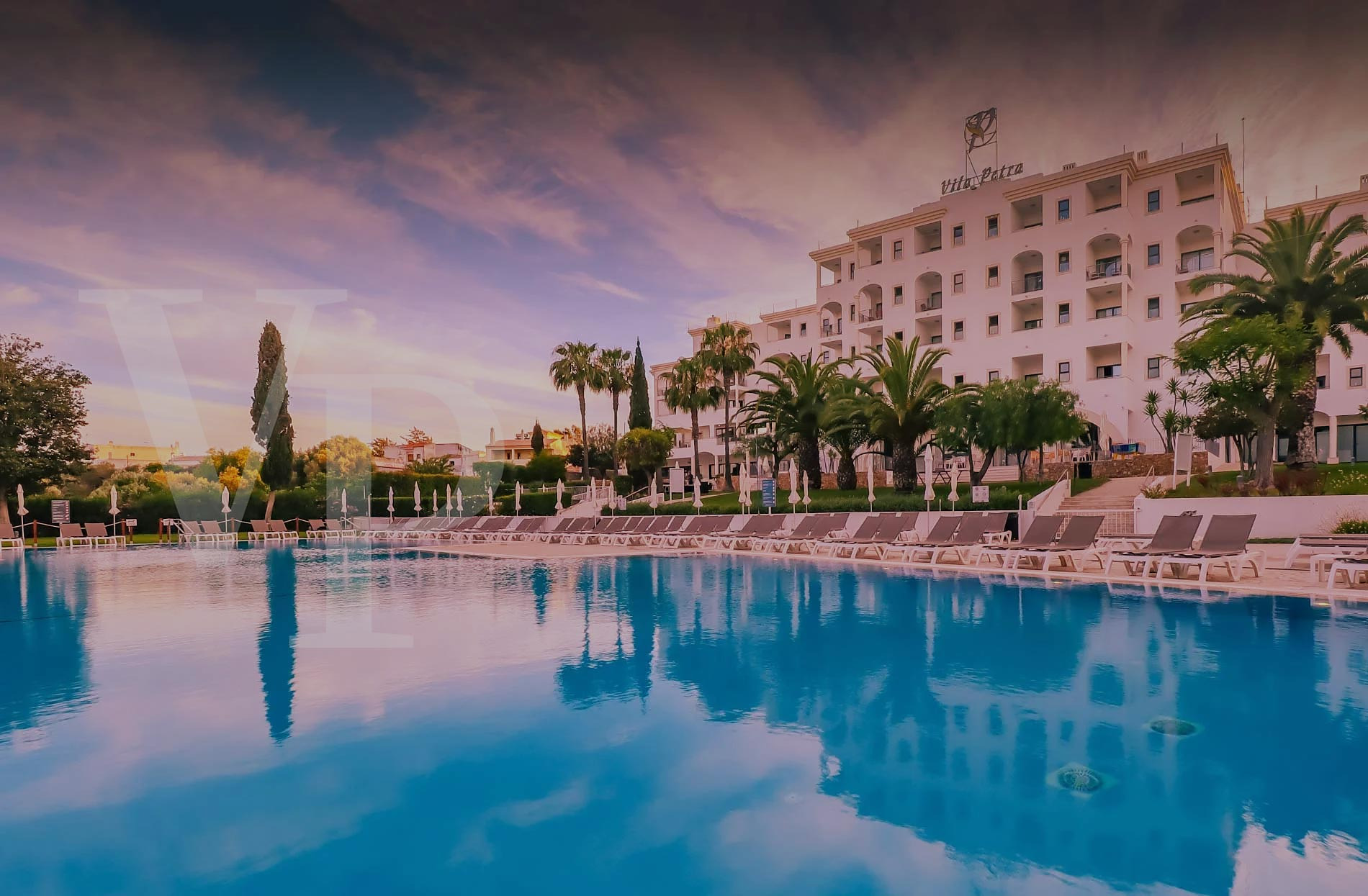 Piscina e hotel Vila Petra ao pôr do sol, com espreguiçadeiras e palmeiras.