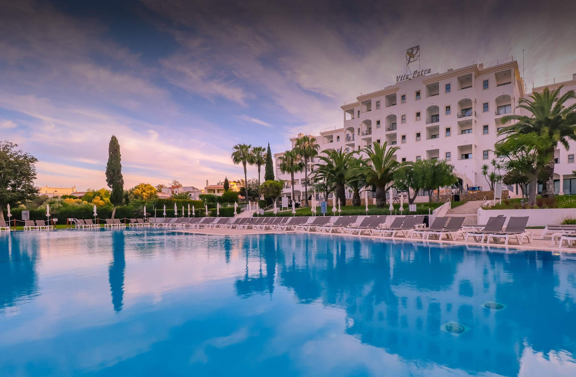 Piscina e hotel Vila Petra ao pôr do sol, com espreguiçadeiras e palmeiras.