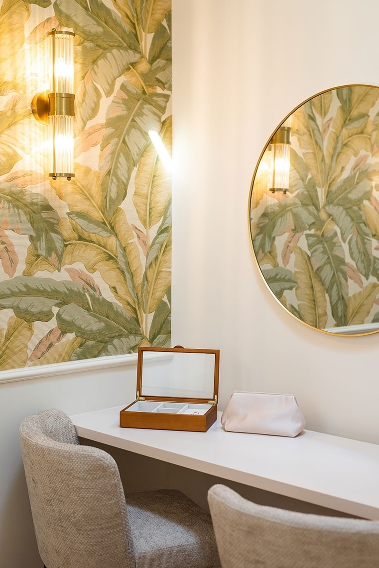 Área de vaidade de hotel com papel de parede tropical, espelho redondo e luminárias elegantes.