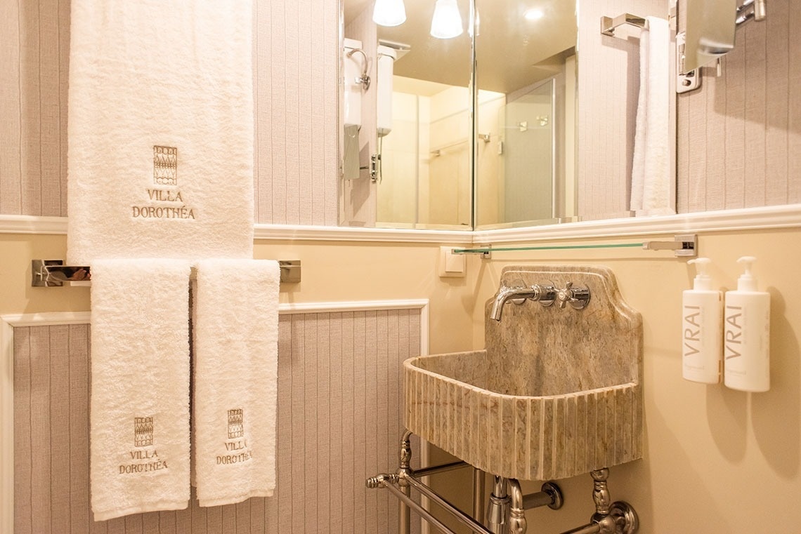 Banheiro elegante de hotel com pia de mármore, toalhas bordadas Villa Dorothéa e produtos de higiene.
