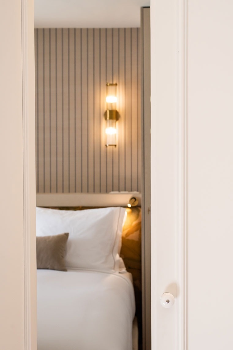 Quarto de hotel elegante com cama, travesseiros, parede listrada e arandela dourada, visto da porta.