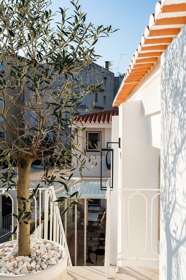This image captures a bright outdoor patio area featuring white buildings with balconies, a wooden pergola covering outdoor seating, and a prominent spiral staircase in the background.