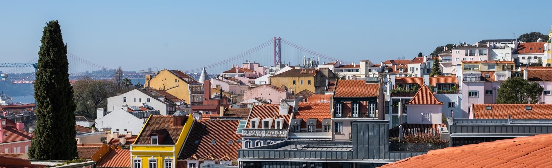 Um pátio ensolarado com pérgula de madeira e área de refeições, rodeado por edifícios brancos com varandas e uma escada em caracol.