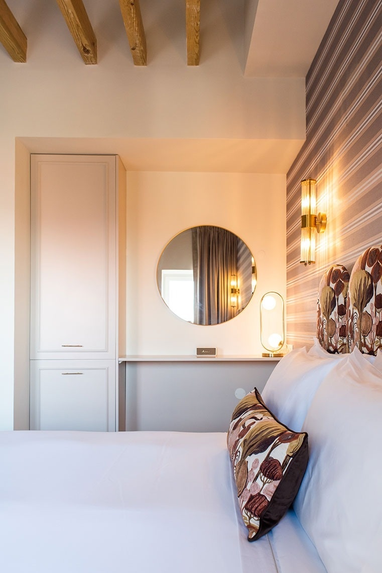 Quarto de hotel elegante: cama branca, almofada estampada, espelho, luminárias douradas e vigas de madeira.