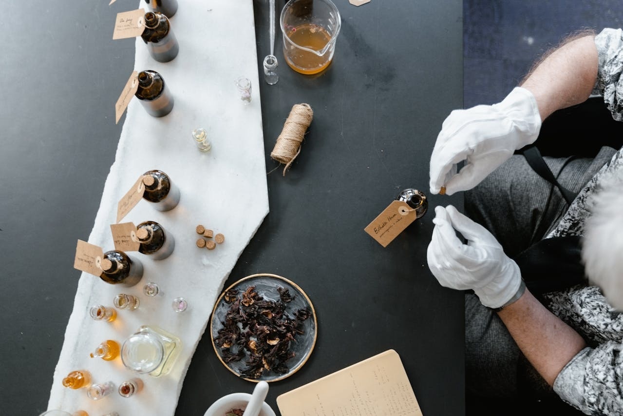 An overhead shot captures a person wearing white gloves carefully placing an ingredient into a small dark bottle, surrounded by numerous other vials, dried botanicals, and crafting supplies on a dark table.