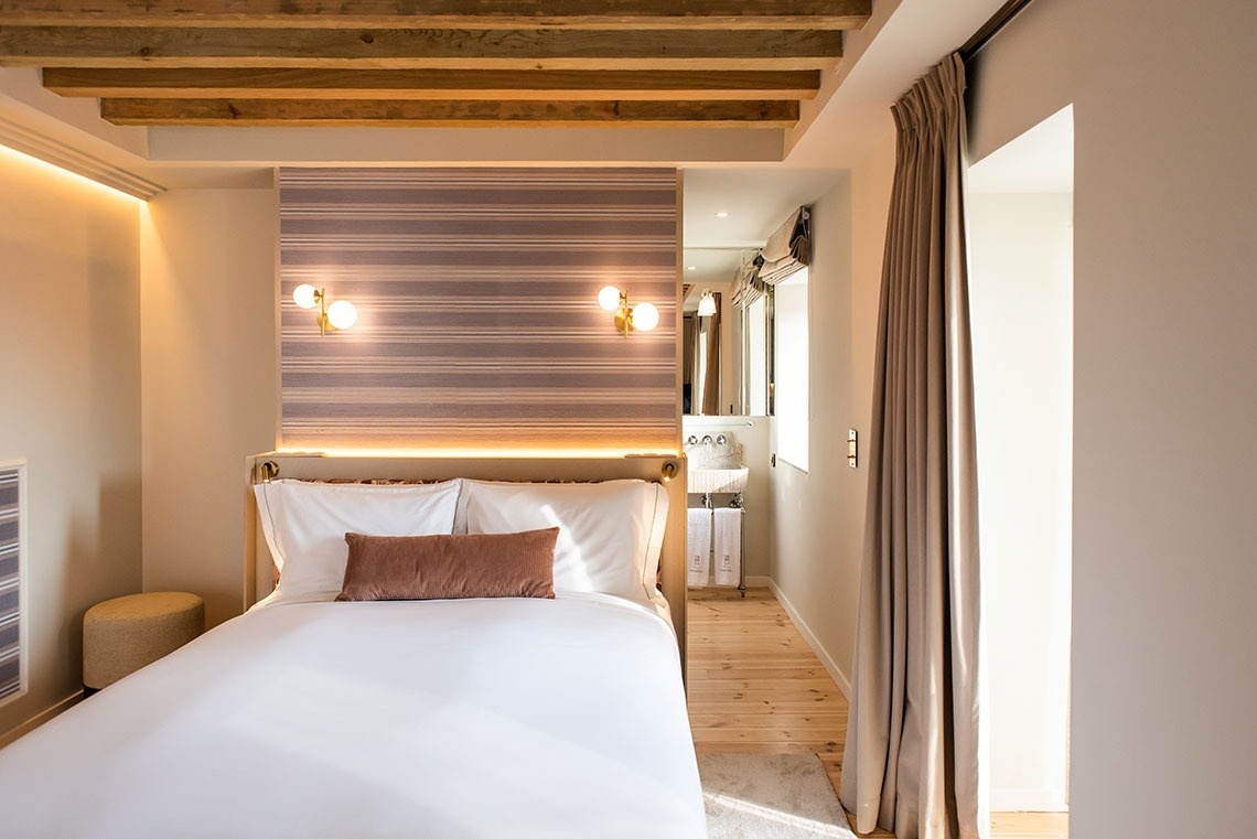 Quarto de hotel aconchegante com cama confortável, parede listrada, vigas de madeira e iluminação ambiente.