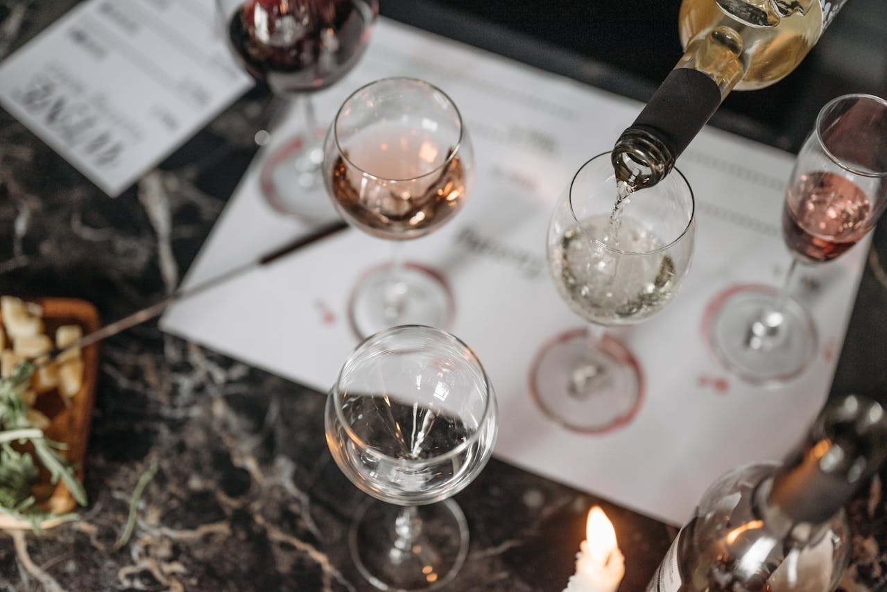 White wine is poured from a bottle into a glass, surrounded by other wine glasses with various wines, snacks, and a lit candle on a dark marble surface, suggesting a wine tasting or gathering.