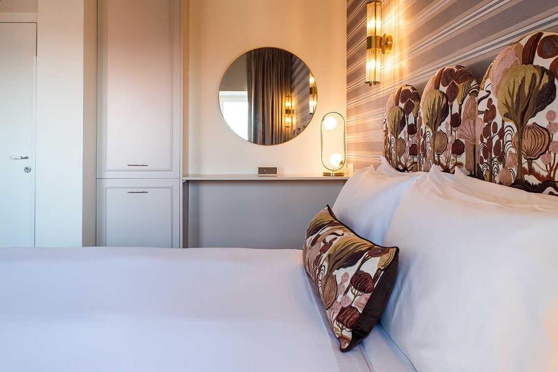 Quarto de hotel moderno com cama, cabeceira de padrão botânico, espelho e iluminação ambiente. Conforto e estilo.