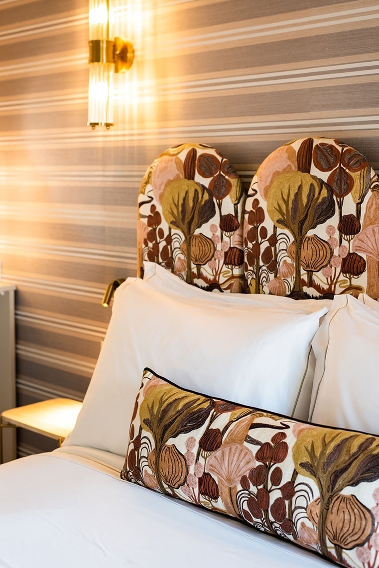 Cama de hotel com cabeceira e almofada de estampa artística, iluminação quente e papel de parede.