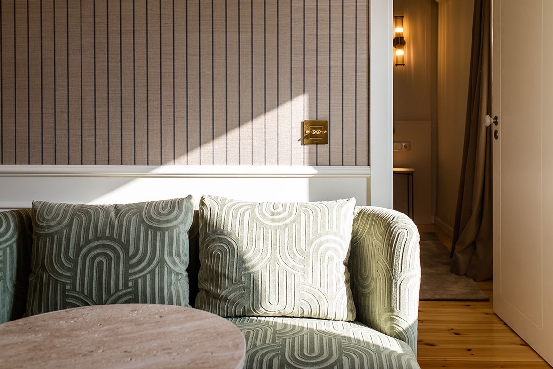 Quarto de hotel acolhedor com sofá verde, almofadas estampadas, papel de parede listrado e luz natural.