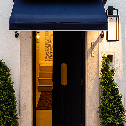 Entrada de la Villa Dorothea: puerta negra, toldo azul y olivos en maceta.