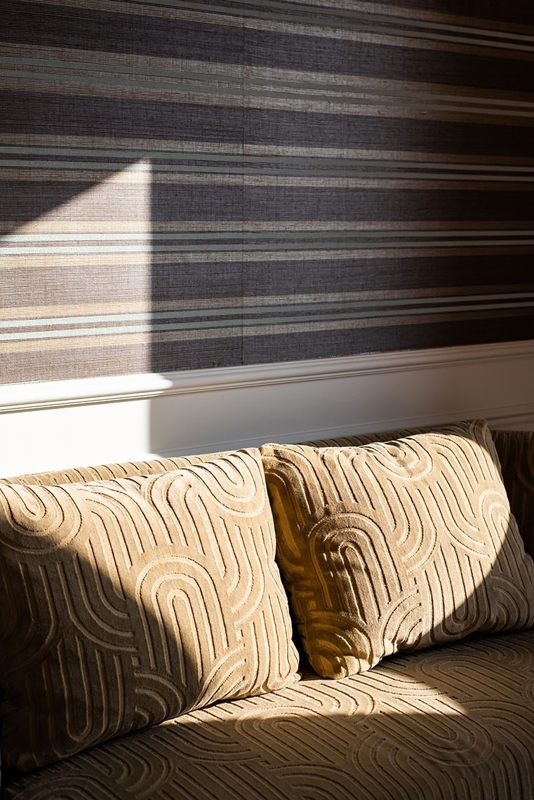 Confortável sofá de hotel com almofadas estampadas, parede listrada e luz natural.