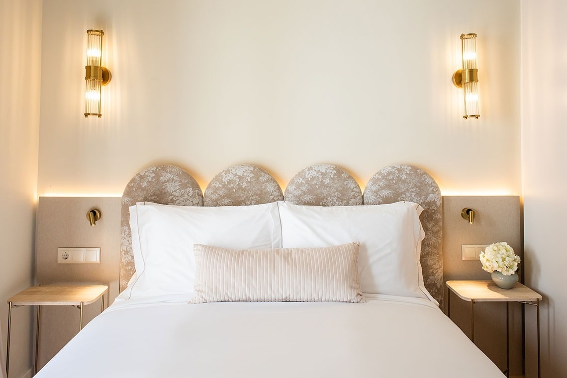 Quarto de hotel elegante com cama de casal, cabeceira floral, luminárias douradas e iluminação ambiente acolhedora.