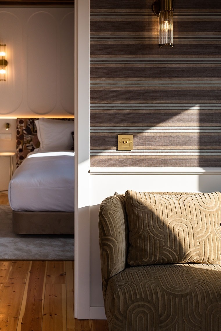 Quarto de hotel moderno com poltrona texturizada, parede listrada e cama aconchegante, sob luz natural.