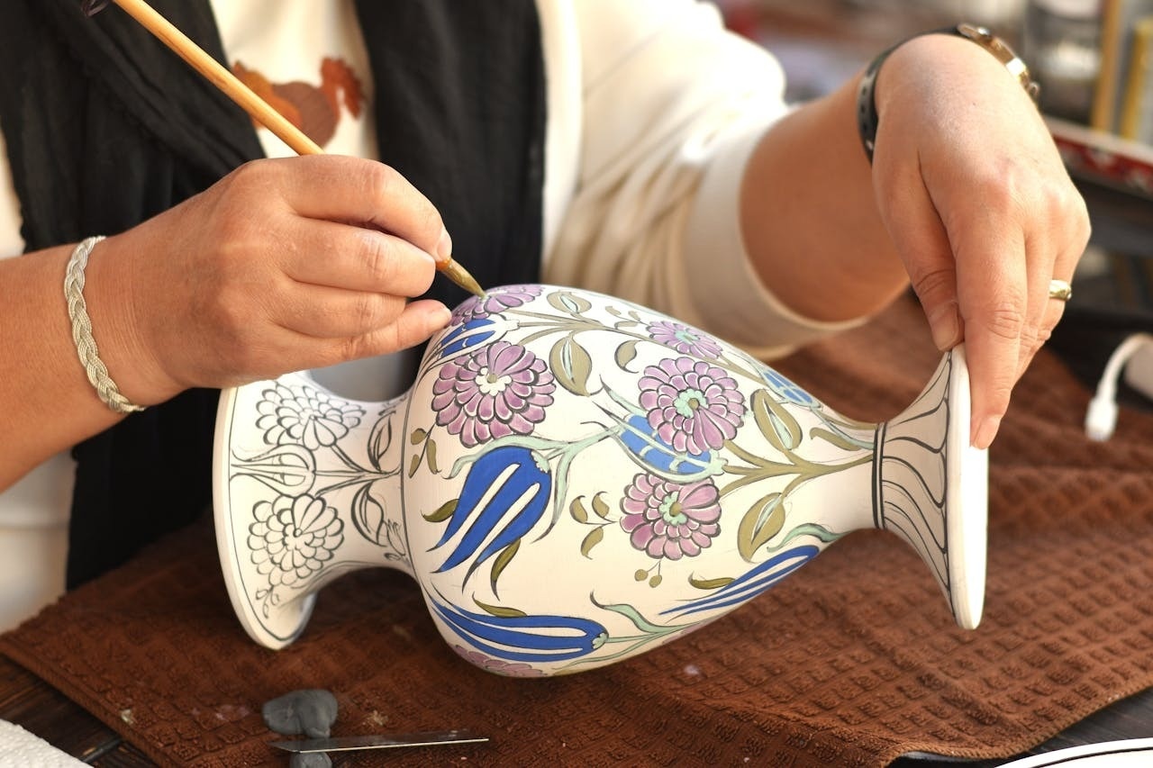A person's hands are meticulously painting intricate floral designs onto a ceramic vase with a fine brush.