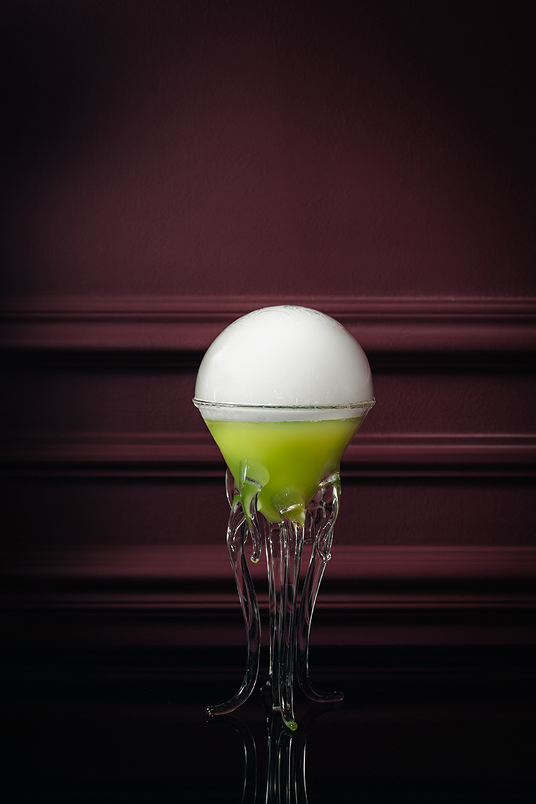 A spooky green cocktail with a frothy top, served in a unique glass Villa Dorothea, in a dimly lit bar.