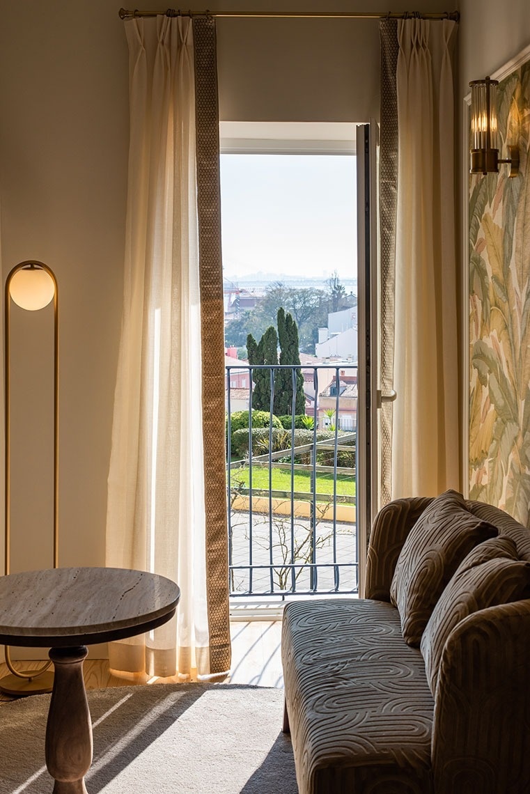 Quarto de hotel luminoso com varanda, vista cidade e jardim, e sofá convidativo.