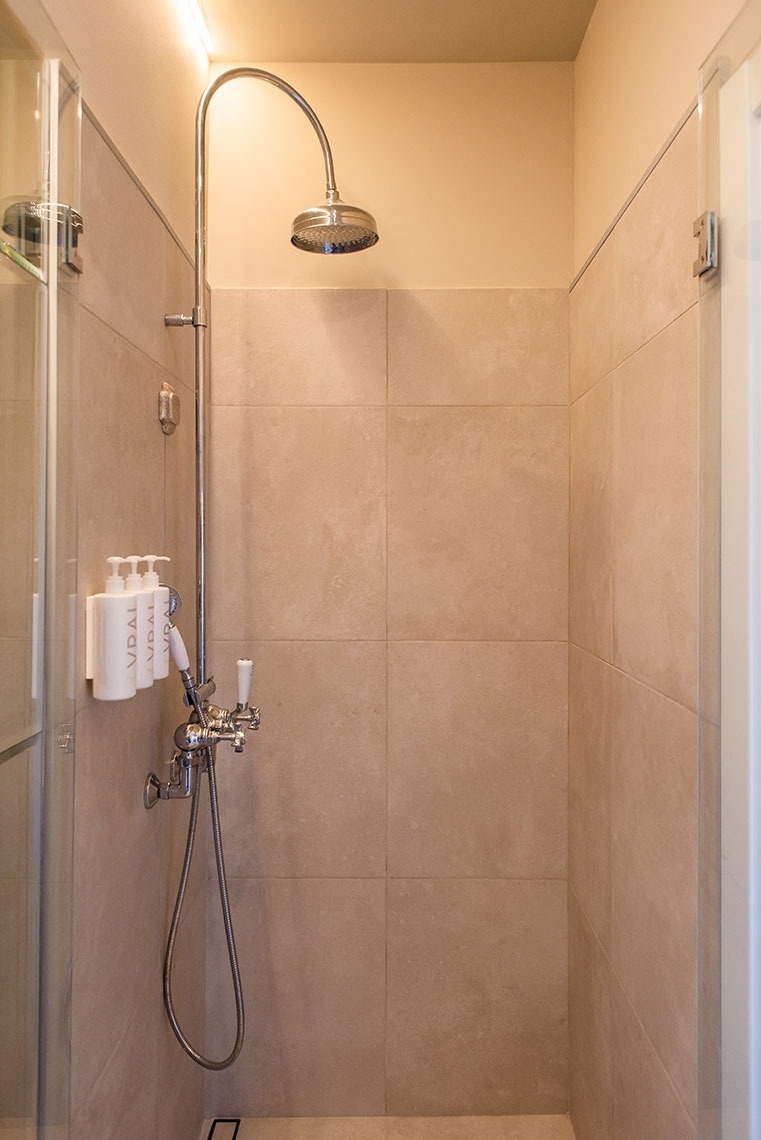 Chuveiro moderno de hotel com azulejos em tons neutros, chuveiro de teto e de mão, e dispensadores de amenities.