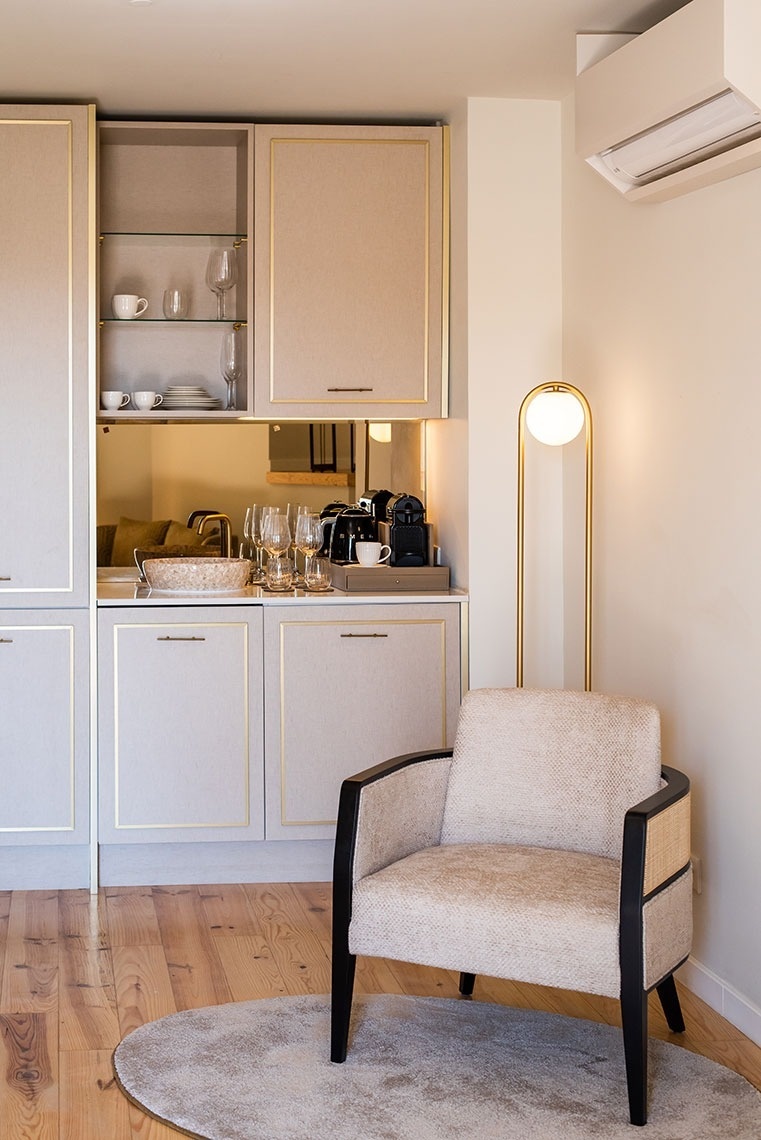 Quarto de hotel com kitchenette equipada, poltrona aconchegante e luminária dourada.