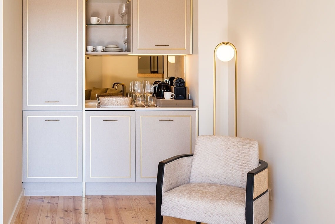 Minibar e estação de café de luxo em quarto de hotel com poltrona confortável e luminária dourada.