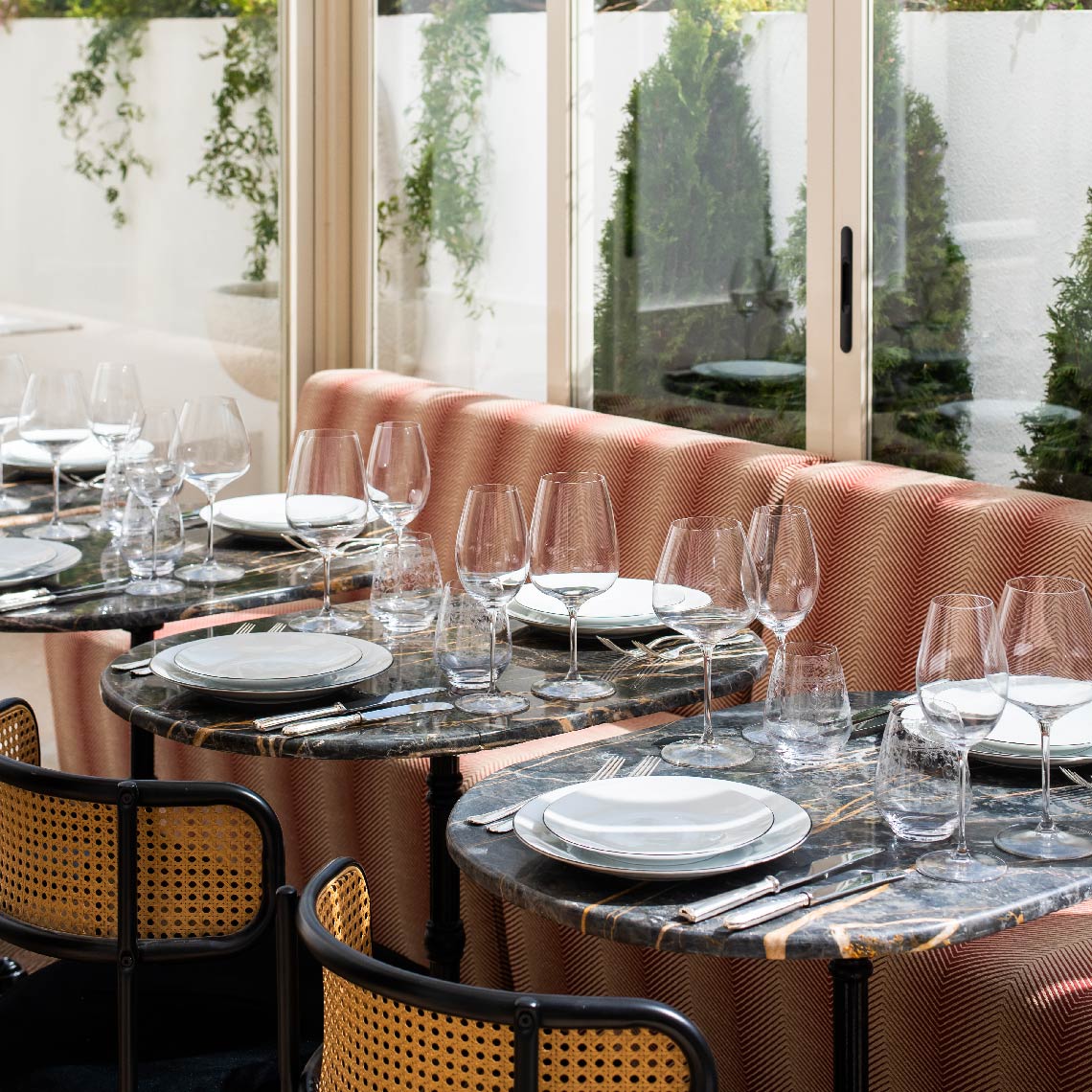 Mesa de restaurante lista para Villa Dorothea con copas y vajilla sobre mesas de mármol y asientos tapizados.
