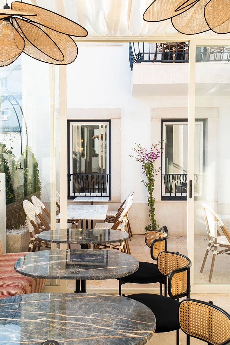 An architectural rendering depicts a sunlit outdoor patio with a wooden pergola and dining set, surrounded by white multi-story buildings featuring balconies, windows with stone frames, and a spiral staircase.